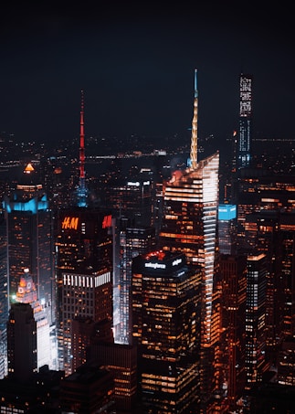 A nighttime cityscape featuring illuminated skyscrapers with distinct architectural designs. The buildings display vibrant lights and advertisements from companies like H&M and Salesforce. The atmosphere is urban and bustling, with a dark sky providing contrast to the bright city lights.