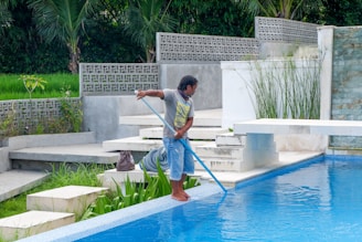 A person is cleaning a swimming pool using a long pool skimmer. They are dressed in casual attire and standing barefoot on the pool's edge. The surroundings include lush green plants, large potted plants, and decorative concrete walls. The scene conveys a peaceful and serene outdoor environment.