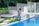 A person is cleaning a swimming pool using a long pool skimmer. They are dressed in casual attire and standing barefoot on the pool's edge. The surroundings include lush green plants, large potted plants, and decorative concrete walls. The scene conveys a peaceful and serene outdoor environment.