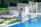 A person is cleaning a swimming pool using a long pool skimmer. They are dressed in casual attire and standing barefoot on the pool's edge. The surroundings include lush green plants, large potted plants, and decorative concrete walls. The scene conveys a peaceful and serene outdoor environment.