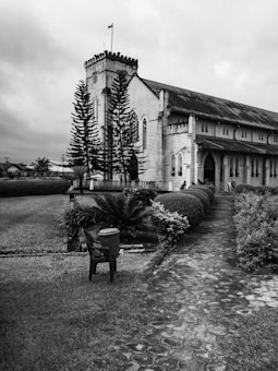 A historic-looking church made of stone, featuring a prominent tower and arched windows. Lush landscaping with neatly trimmed bushes and large conifer trees surrounds the path leading up to the entrance. A solitary plastic chair with a container placed on it is situated on the grass beside the path.
