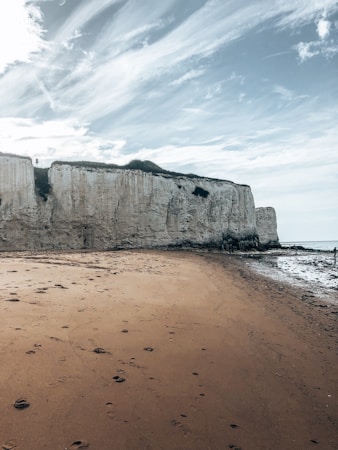 A rugged coastline featuring tall, white chalk cliffs with grass on top. The foreground shows a sandy beach with footprints leading toward the water. The sky is partly cloudy with streaks of white clouds against a blue backdrop.