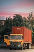 A yellow Tata truck is parked with a large red shipping container on its trailer. The container has the word 'PERMA' printed on it. The truck's driver-side door is open, and it's positioned in front of lush green trees and under a vibrant pink and blue sky.