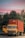 A yellow Tata truck is parked with a large red shipping container on its trailer. The container has the word 'PERMA' printed on it. The truck's driver-side door is open, and it's positioned in front of lush green trees and under a vibrant pink and blue sky.