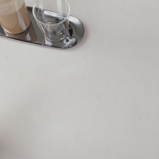 A minimalistic scene featuring a reflective metallic tray on a light surface. On the tray, there is a glass filled with a creamy beverage next to an empty glass.