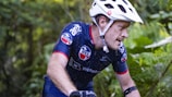 A cyclist wearing a white helmet and a navy blue cycling jersey is intensely focused as he rides through a lush, green forest. His jersey is adorned with several logos, and he appears to be in motion, with small droplets of water visible on his skin. The environment is dense with greenery, suggesting an off-road trail or a mountain biking path.