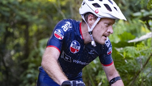 A cyclist wearing a white helmet and a navy blue cycling jersey is intensely focused as he rides through a lush, green forest. His jersey is adorned with several logos, and he appears to be in motion, with small droplets of water visible on his skin. The environment is dense with greenery, suggesting an off-road trail or a mountain biking path.