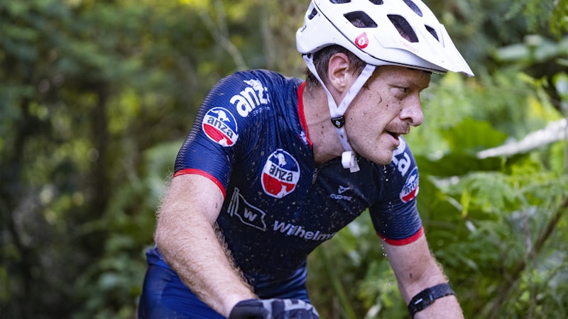 A cyclist wearing a white helmet and a navy blue cycling jersey is intensely focused as he rides through a lush, green forest. His jersey is adorned with several logos, and he appears to be in motion, with small droplets of water visible on his skin. The environment is dense with greenery, suggesting an off-road trail or a mountain biking path.