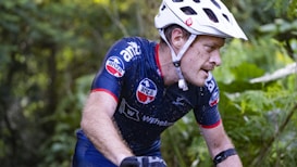 A cyclist wearing a white helmet and a navy blue cycling jersey is intensely focused as he rides through a lush, green forest. His jersey is adorned with several logos, and he appears to be in motion, with small droplets of water visible on his skin. The environment is dense with greenery, suggesting an off-road trail or a mountain biking path.