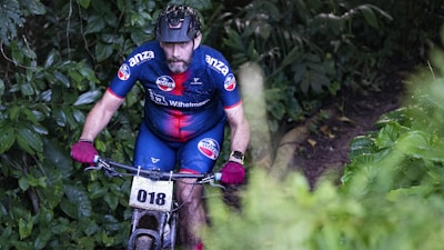 A cyclist wearing a blue and red racing suit with a number 018 attached to the bicycle rides through a dense forest trail. The cyclist is wearing a helmet and gloves, and the surrounding area is lush with green foliage.