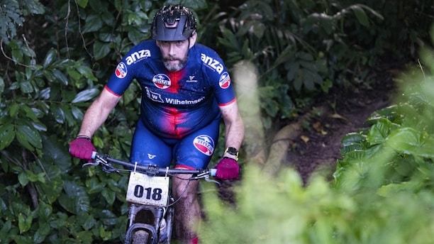 A cyclist wearing a blue and red racing suit with a number 018 attached to the bicycle rides through a dense forest trail. The cyclist is wearing a helmet and gloves, and the surrounding area is lush with green foliage.