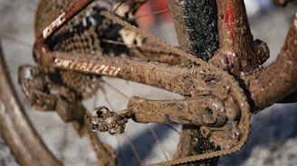 A close-up view of a bicycle's drivetrain covered with thick mud, showing the chain, pedals, and part of the frame. The mud is caked onto the components, indicating the bicycle has been ridden in a rough, wet environment.
