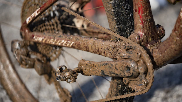 A close-up view of a bicycle's drivetrain covered with thick mud, showing the chain, pedals, and part of the frame. The mud is caked onto the components, indicating the bicycle has been ridden in a rough, wet environment.