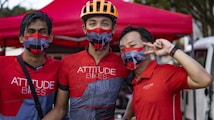 Three individuals wearing red and gray patterned face masks are standing closely together, appearing cheerful and united. They are dressed in cycling jerseys and shirts with the slogan 'ATTITUDE BIKES' printed on them. A bright orange helmet is worn by the person in the middle. The group is positioned in front of a red tent with a relaxed outdoor setting.
