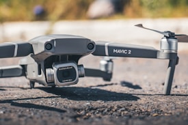 A Mavic 2 drone is positioned on the ground, showcasing its camera labeled with 'HASSELBLAD'. The background is slightly blurred, highlighting the drone's sleek design and metallic finish.