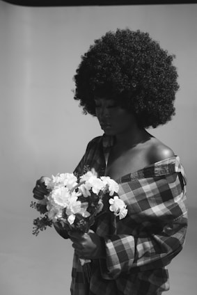 A person with a large afro hairstyle is gently holding a bouquet of white flowers while wearing a plaid shirt with an open neckline. The scene is in black and white, and the subject appears to be looking down at the flowers.