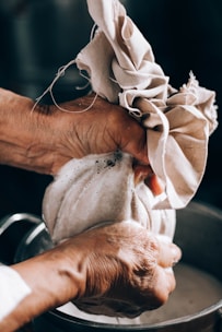 A pair of hands squeezes a cloth bag over a container, with liquid seeping through the cloth. The hands appear aged and the cloth is wrinkled, suggesting a manual process, possibly related to cooking or cheese-making.