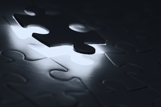 A single puzzle piece is slightly raised above its incomplete space, surrounded by other interlocking pieces. Soft, diffused lighting emanates from beneath, creating a dramatic contrast between light and shadow.