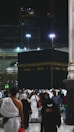 A large gathering of people surrounds the Kaaba at night, illuminated by artificial lights. Many individuals are dressed in traditional pilgrimage attire. The backdrop includes construction cranes and partially lit modern structures.