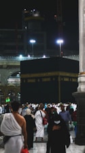 A large gathering of people surrounds the Kaaba at night, illuminated by artificial lights. Many individuals are dressed in traditional pilgrimage attire. The backdrop includes construction cranes and partially lit modern structures.