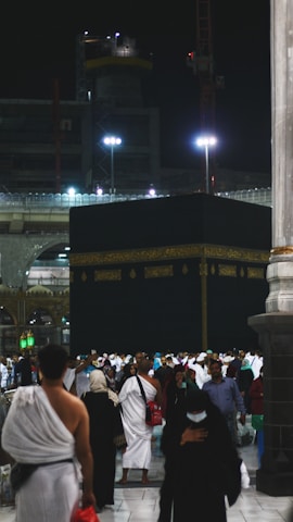 A large gathering of people surrounds the Kaaba at night, illuminated by artificial lights. Many individuals are dressed in traditional pilgrimage attire. The backdrop includes construction cranes and partially lit modern structures.