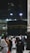 A large gathering of people surrounds the Kaaba at night, illuminated by artificial lights. Many individuals are dressed in traditional pilgrimage attire. The backdrop includes construction cranes and partially lit modern structures.