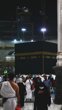 A large gathering of people surrounds the Kaaba at night, illuminated by artificial lights. Many individuals are dressed in traditional pilgrimage attire. The backdrop includes construction cranes and partially lit modern structures.