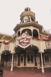 A vintage-style ice cream parlor with decorative pumpkins and autumn-themed garlands. The ornate architecture features a tower with festive Halloween decorations, and the sign reads 'The Gibson Girl Ice Cream Parlour'. The facade is adorned with intricate details, pillars, and a small seating area with white wrought iron chairs and tables.