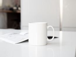 A white ceramic mug with a simple handle is placed on a clean, white surface. An open magazine or brochure lies nearby, partially visible. The background is softly blurred, featuring a light, minimalistic interior with a hint of furniture, suggesting a calm, indoor setting.