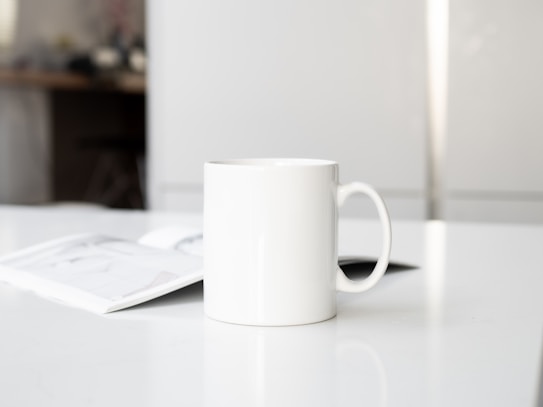 A white ceramic mug with a simple handle is placed on a clean, white surface. An open magazine or brochure lies nearby, partially visible. The background is softly blurred, featuring a light, minimalistic interior with a hint of furniture, suggesting a calm, indoor setting.