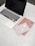 A laptop with a black screen sits on a white marble surface. To the right of the laptop, there are a pair of wireless earbuds placed on top of a light pink notebook. Underneath the notebook is a red notebook with small patterns. Nearby, a pair of round eyeglasses rests on the red notebook.