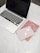 A laptop with a black screen sits on a white marble surface. To the right of the laptop, there are a pair of wireless earbuds placed on top of a light pink notebook. Underneath the notebook is a red notebook with small patterns. Nearby, a pair of round eyeglasses rests on the red notebook.