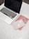 A laptop with a black screen sits on a white marble surface. To the right of the laptop, there are a pair of wireless earbuds placed on top of a light pink notebook. Underneath the notebook is a red notebook with small patterns. Nearby, a pair of round eyeglasses rests on the red notebook.