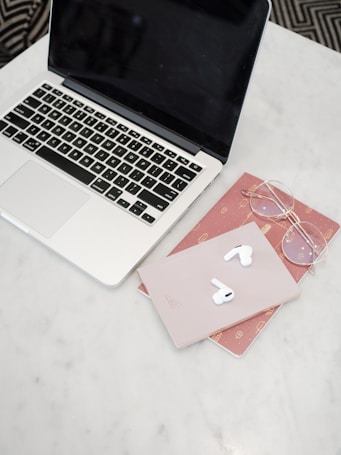 A laptop with a black screen sits on a white marble surface. To the right of the laptop, there are a pair of wireless earbuds placed on top of a light pink notebook. Underneath the notebook is a red notebook with small patterns. Nearby, a pair of round eyeglasses rests on the red notebook.