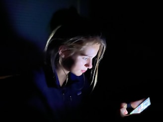 A young person is illuminated by the light of a smartphone screen, which they are intently looking at in a dimly lit environment. Their face is partially visible, and the surrounding area is in shadow.
