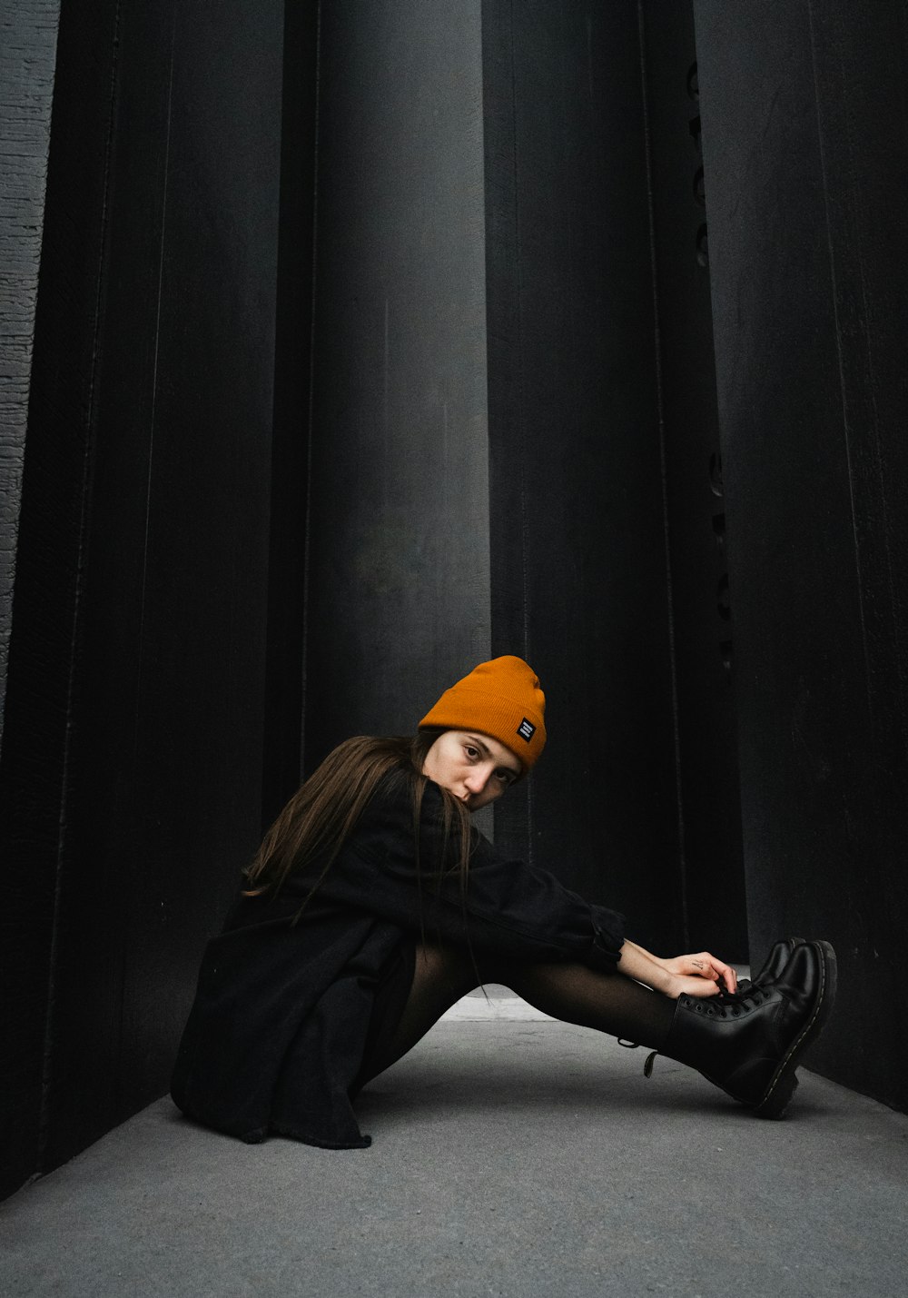 woman in black leather jacket and orange knit cap sitting on black wooden wall