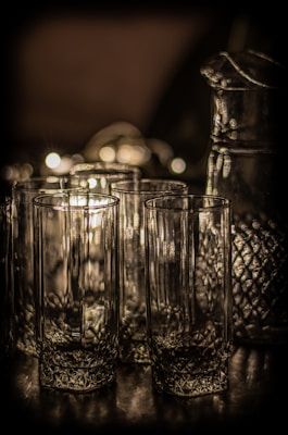 A collection of intricately designed glassware, including tall glasses and a matching pitcher, with a textured surface that reflects light in a dim setting. The glasses have a star-like pattern at the base and stand on a reflective surface, creating subtle highlights and shadows.