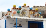A building facade featuring a rustic, nautical theme. The roof is decorated with colorful buoy ornaments in blue, yellow, orange, and green. A life preserver with the word 'MARIA' hangs prominently. Various ropes and antique nautical items add to the maritime decor. Below the roof, a blue-and-white color scheme is evident with a painted mural and a window beneath an awning.
