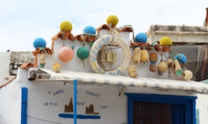 A building facade featuring a rustic, nautical theme. The roof is decorated with colorful buoy ornaments in blue, yellow, orange, and green. A life preserver with the word 'MARIA' hangs prominently. Various ropes and antique nautical items add to the maritime decor. Below the roof, a blue-and-white color scheme is evident with a painted mural and a window beneath an awning.