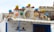 A building facade featuring a rustic, nautical theme. The roof is decorated with colorful buoy ornaments in blue, yellow, orange, and green. A life preserver with the word 'MARIA' hangs prominently. Various ropes and antique nautical items add to the maritime decor. Below the roof, a blue-and-white color scheme is evident with a painted mural and a window beneath an awning.
