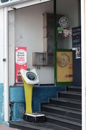 The image features a storefront with a digital weighing scale positioned near the entrance. The scale has a retro design with a yellow stand and a circular display. Surrounding the scale, there are various advertisements, including one for Coca-Cola and another for Heineken. A small staircase leads into the store, and there are signs on the door indicating instructions in Spanish, suggesting a casual urban setting.