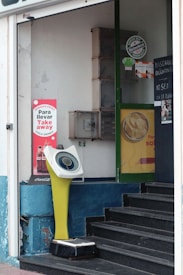 The image features a storefront with a digital weighing scale positioned near the entrance. The scale has a retro design with a yellow stand and a circular display. Surrounding the scale, there are various advertisements, including one for Coca-Cola and another for Heineken. A small staircase leads into the store, and there are signs on the door indicating instructions in Spanish, suggesting a casual urban setting.