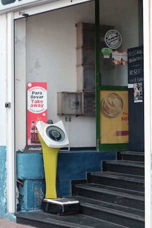 The image features a storefront with a digital weighing scale positioned near the entrance. The scale has a retro design with a yellow stand and a circular display. Surrounding the scale, there are various advertisements, including one for Coca-Cola and another for Heineken. A small staircase leads into the store, and there are signs on the door indicating instructions in Spanish, suggesting a casual urban setting.