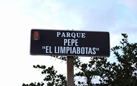 A black and white sign with the words 'PARQUE PEPE EL LIMPIABOTAS' is mounted on a pole against a backdrop of blue sky and green foliage. The top left corner of the sign features a colorful emblem.