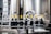 A production line with several wine bottles featuring yellow caps in front of large metal fermentation tanks. The machinery and conveyor belts are part of an industrial setting, and a metal ladder is visible in the background.