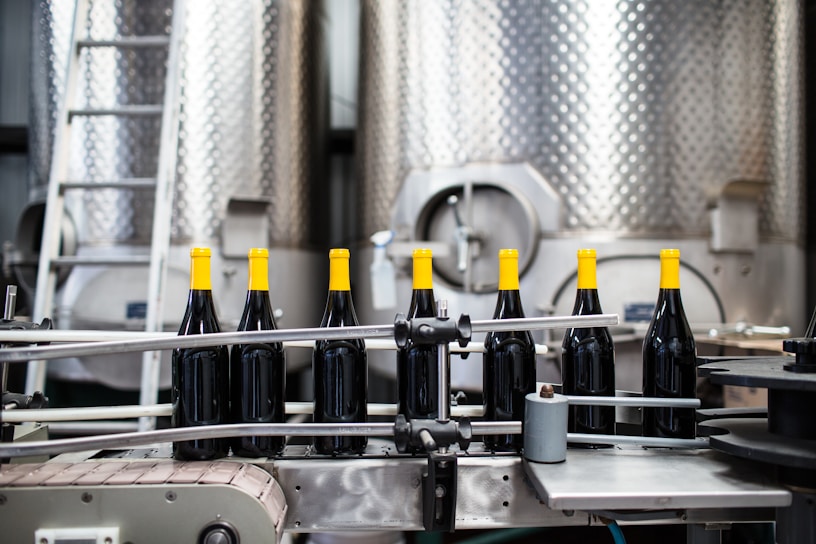A production line with several wine bottles featuring yellow caps in front of large metal fermentation tanks. The machinery and conveyor belts are part of an industrial setting, and a metal ladder is visible in the background.
