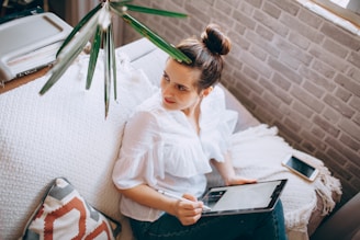 A person is sitting on a couch holding a tablet with a stylus, dressed in a white blouse. The setting includes a light-colored throw and pillow, with a brick wall in the background. A smartphone lies next to them on the couch, under a nearby window filtered by green leaves.