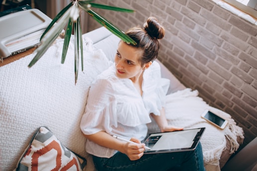 A person is sitting on a couch holding a tablet with a stylus, dressed in a white blouse. The setting includes a light-colored throw and pillow, with a brick wall in the background. A smartphone lies next to them on the couch, under a nearby window filtered by green leaves.