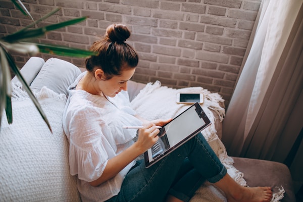 A woman is sitting comfortably on a light-colored sofa with a light blanket draped over it. She is holding a digital tablet and stylus, focusing on writing or drawing. Her hair is tied in a bun, and she is wearing a white blouse and jeans. A smartphone is placed beside her. The environment is calm with natural light coming from a nearby window.