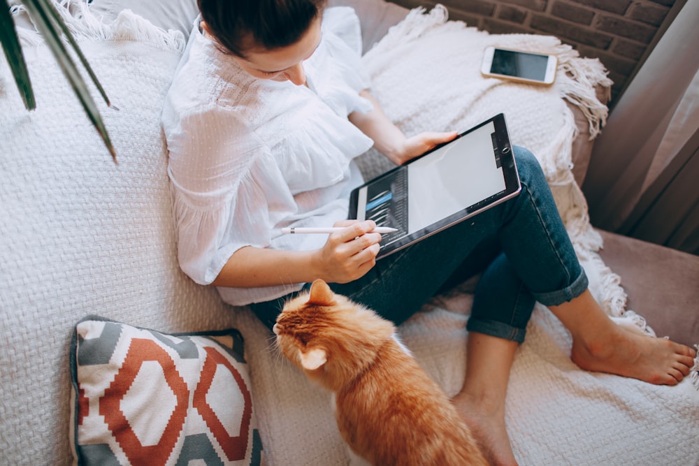 Frau in weißem Langarmhemd und blauen Jeansshorts sitzt mit Tablet-Computer auf dem Bett